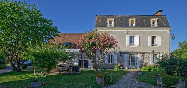A two-story house with a garden, a patio with outdoor furniture, and a clear blue sky. - Le Kouloury (Meyronne)