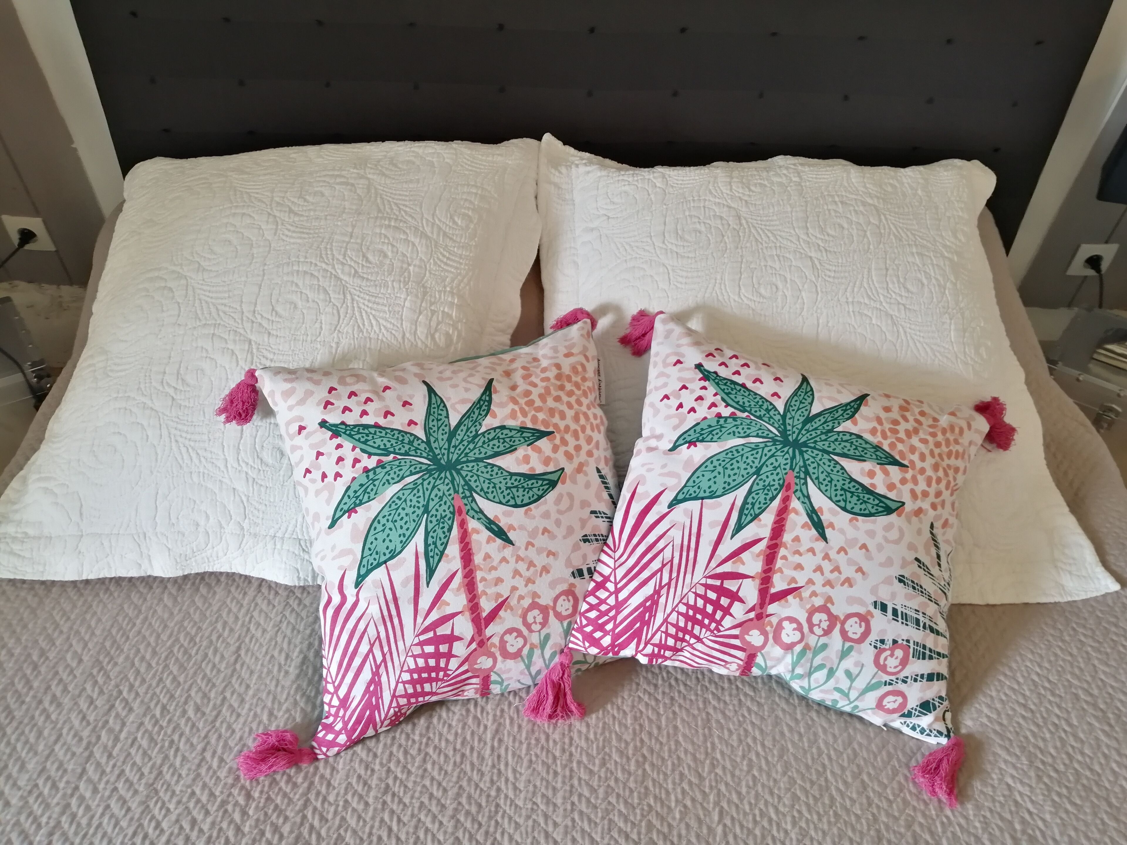 Een bed met twee witte kussens en twee sierkussens met een palmboom- en hartmotief.
