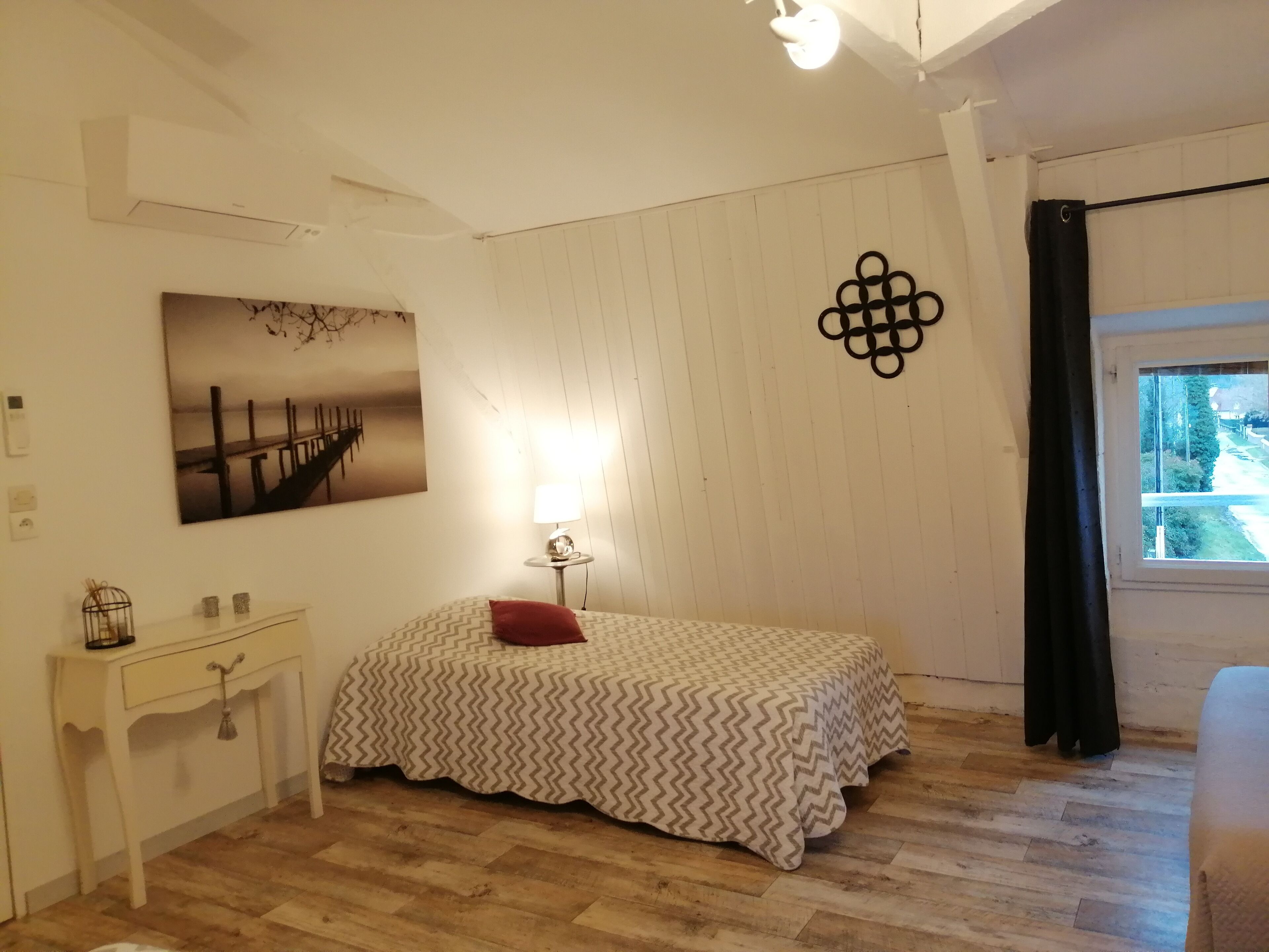 Un dormitorio con cama, mesita de noche, lámpara, un cuadro en la pared y ventana con vistas.