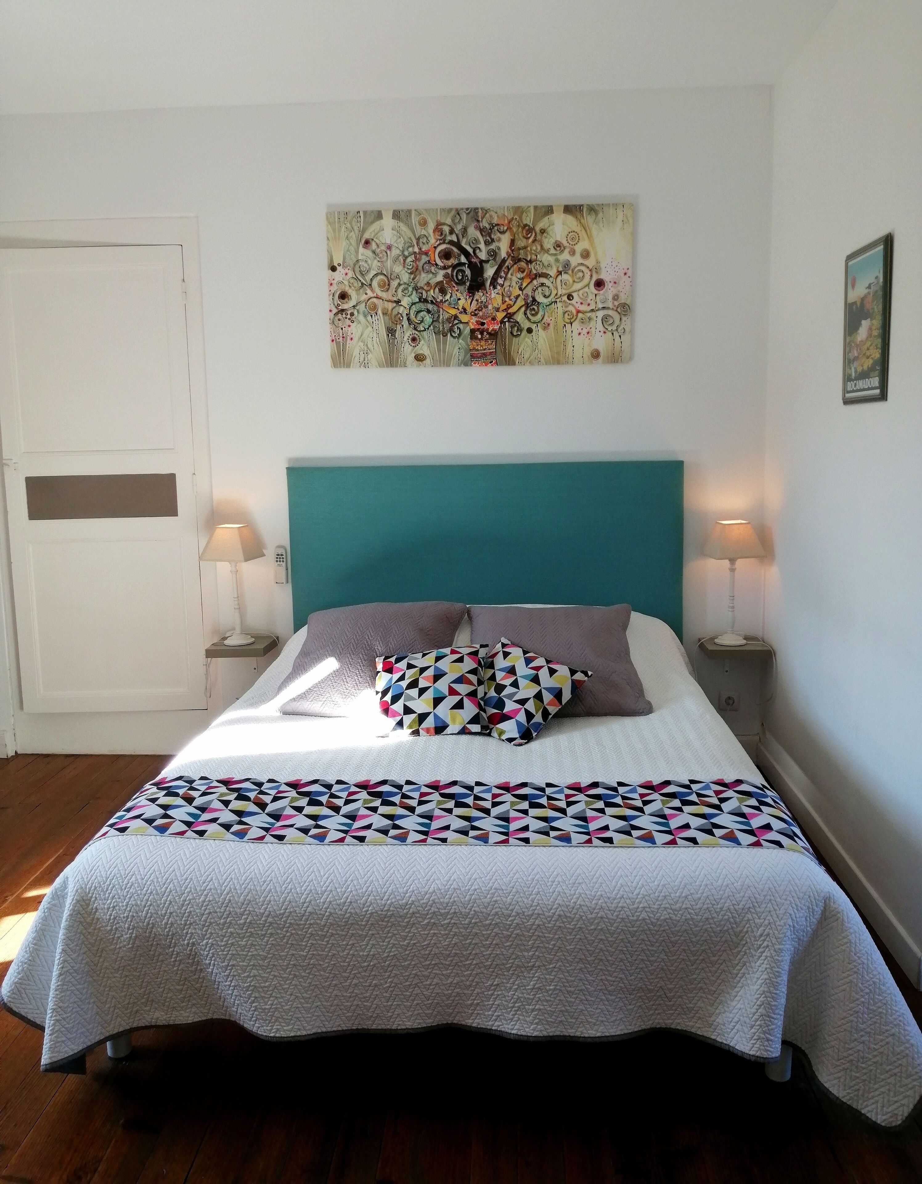 Ein Bett mit petrolfarbenem Kopfteil, zwei Nachttischlampen, einer farbenfrohen geometrischen Decke und einem dekorativen Wandbild darüber.