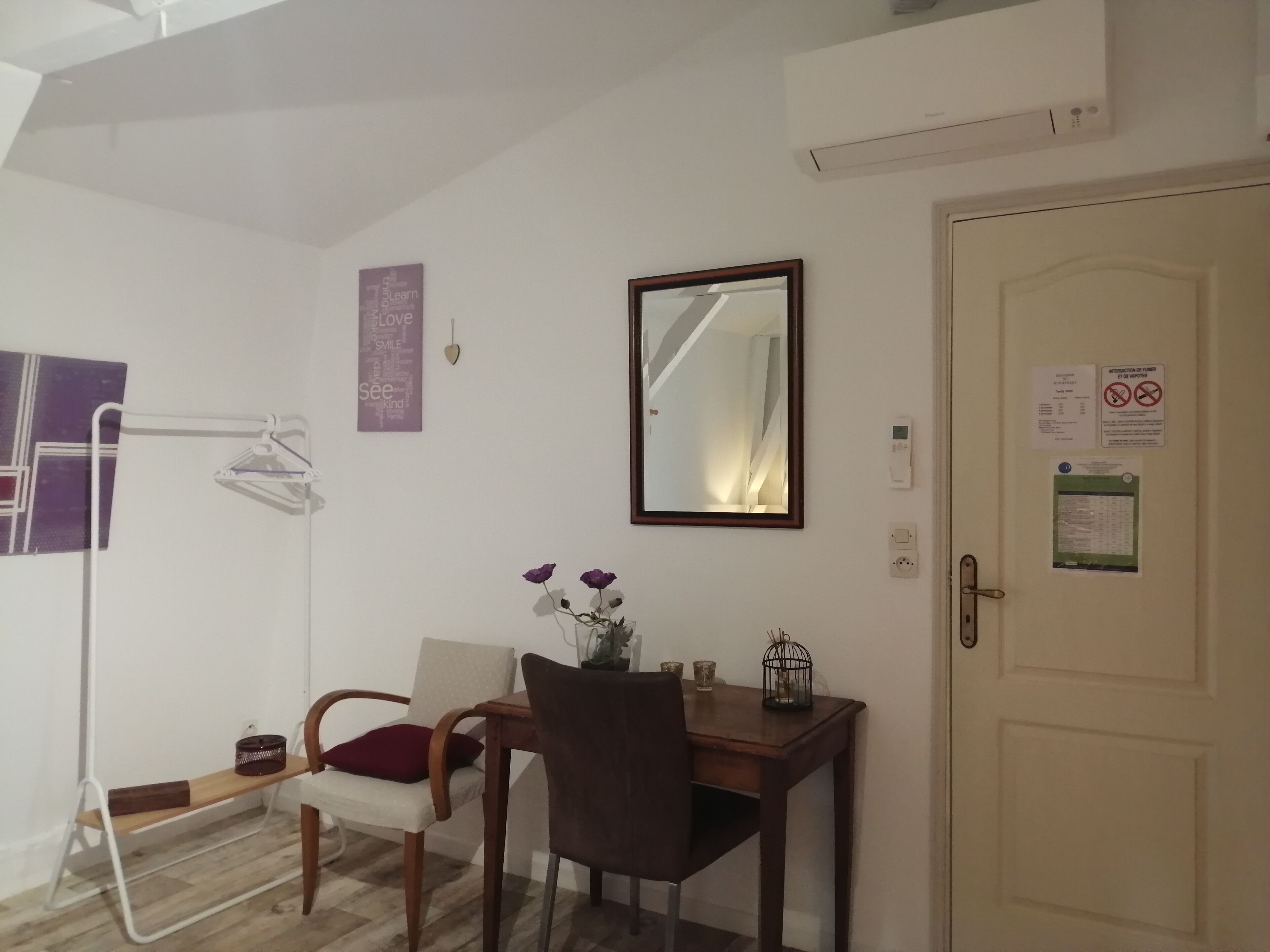Une petite pièce bien éclairée, avec un bureau en bois, une chaise, un miroir et une porte sur laquelle est affichée une annonce.
