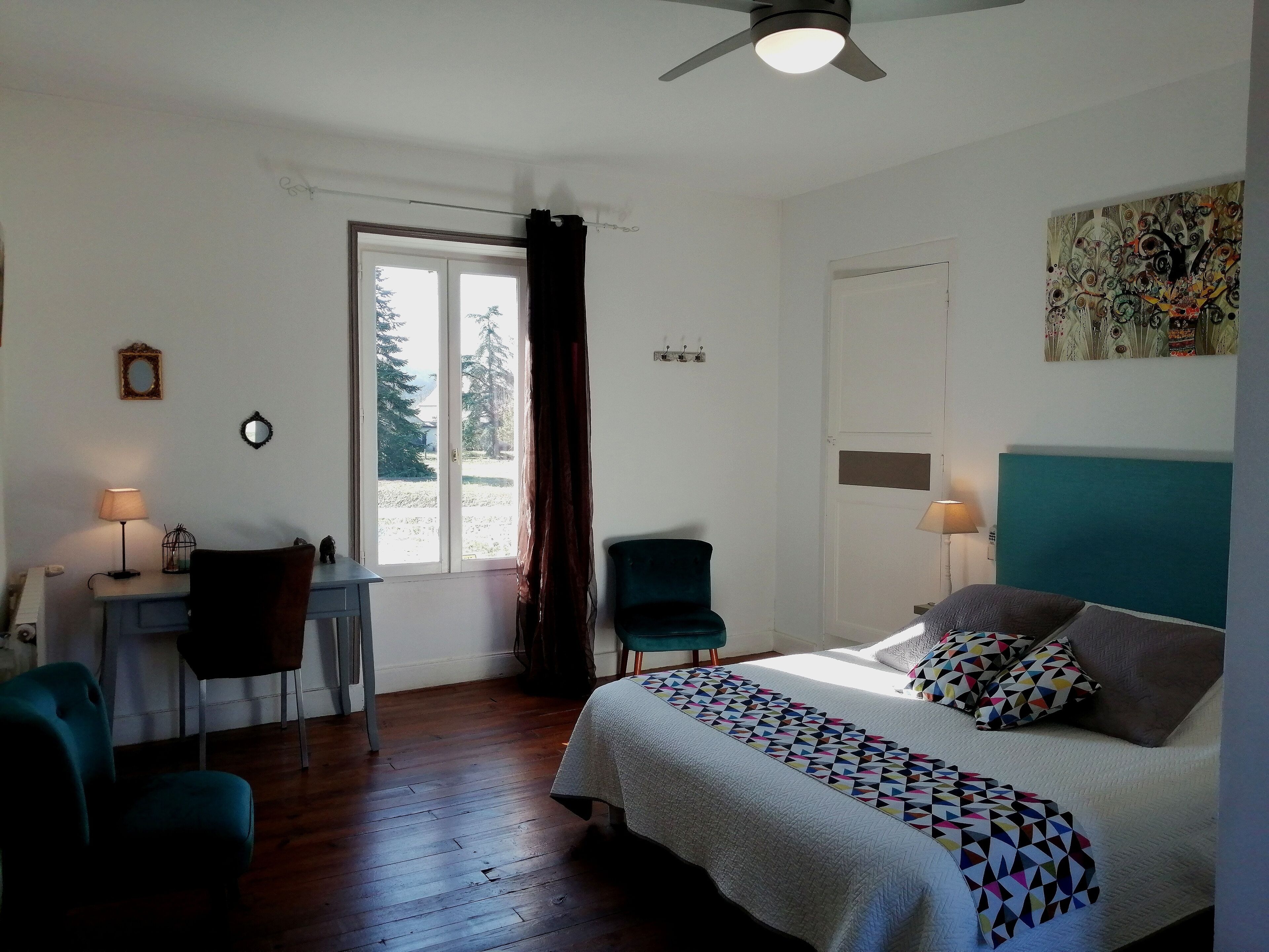 Un dormitorio con cama, escritorio, silla, ventilador de techo y ventana con cortinas.