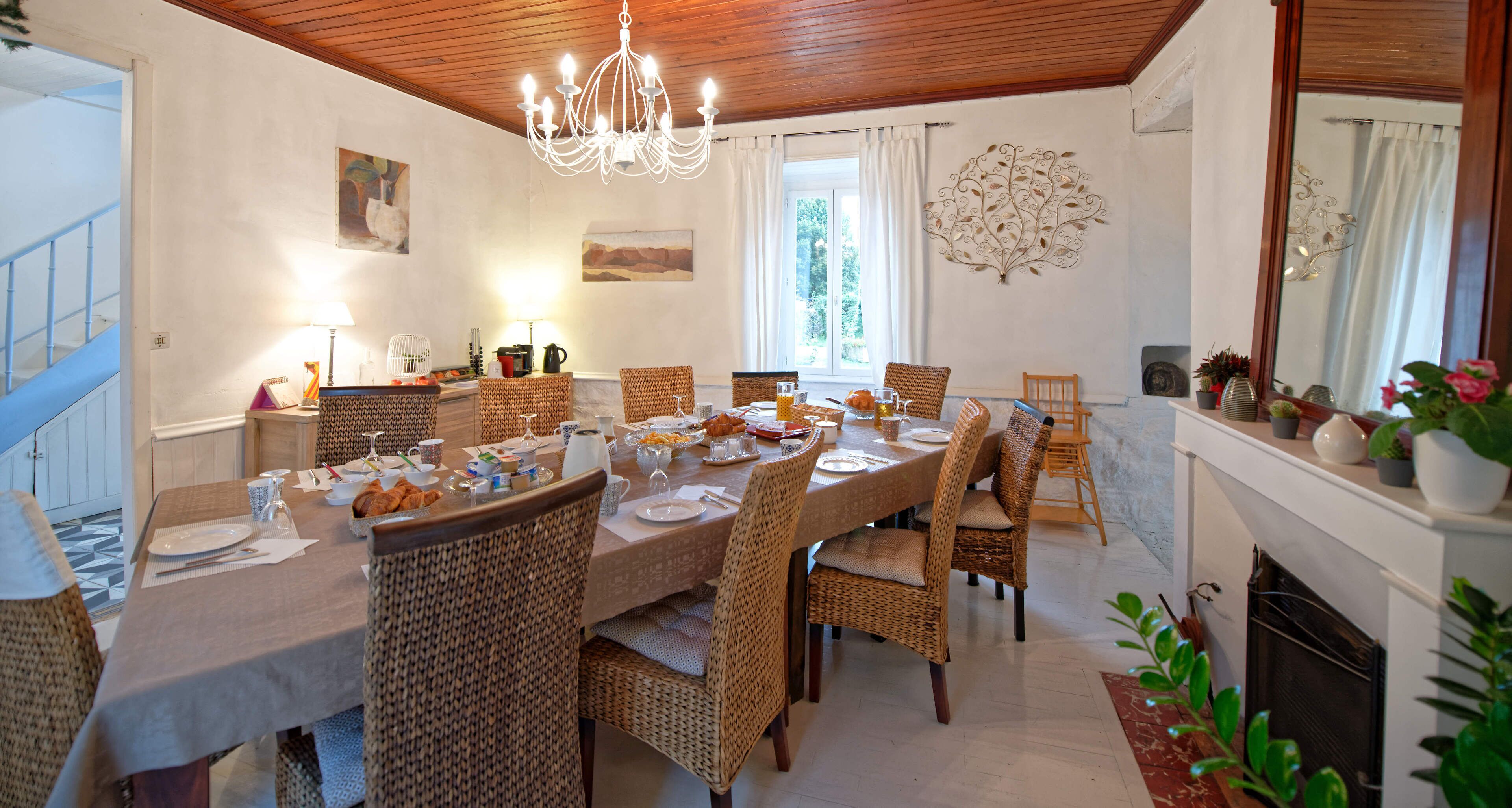A dining room with a long table set for a meal, wicker chairs, a chandelier, and a fireplace.