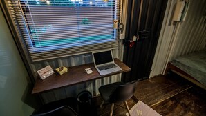 Una habitación pequeña y funcional con un escritorio de madera, una computadora portátil, dos sillas, una ventana con persianas, una puerta con manija y una cama con una manta verde.