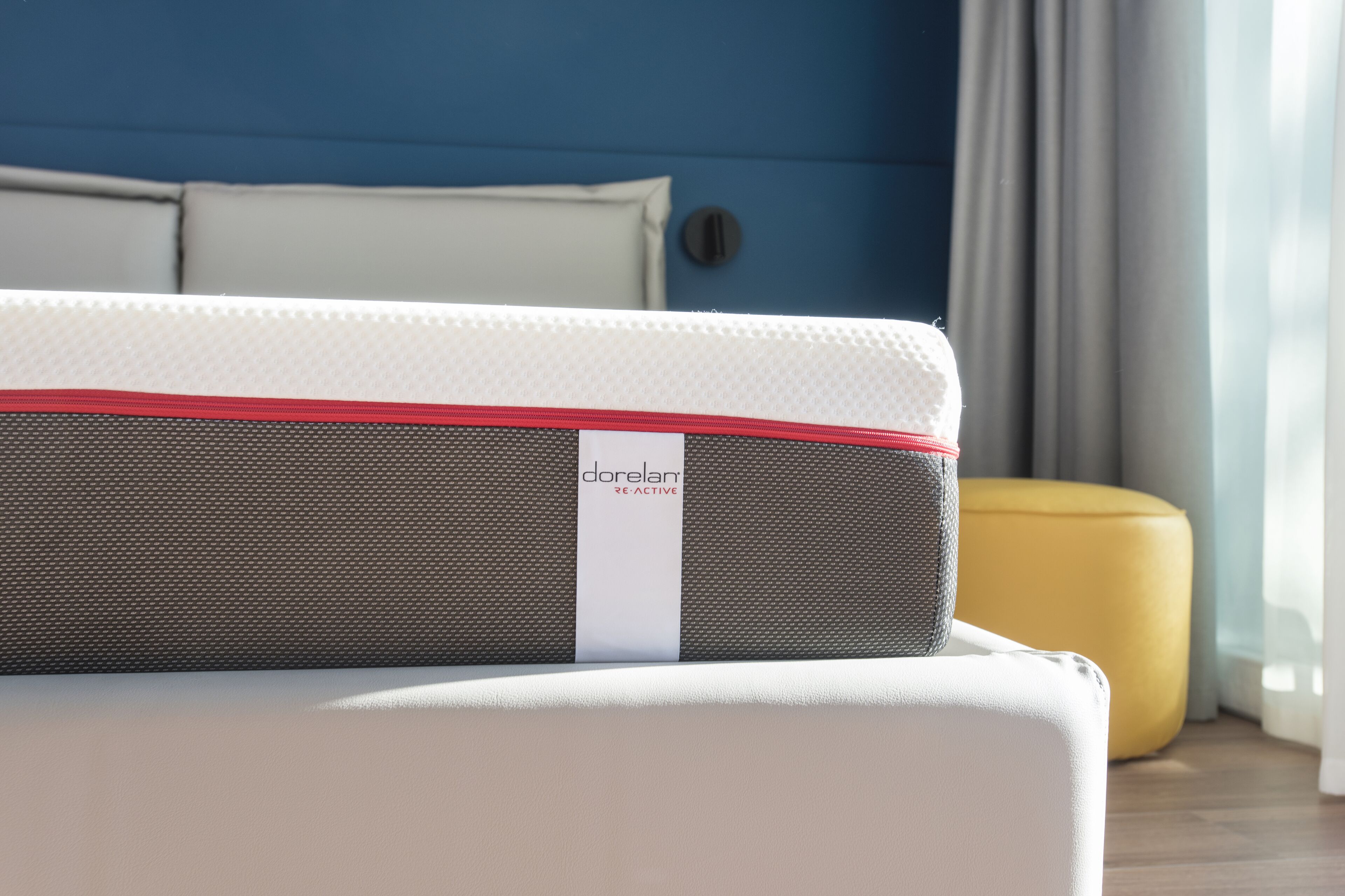 Un sofá blanco con un cojín rojo y gris que dice "dorelam reactive" en una habitación moderna con paredes azules y un otoman amarillo.