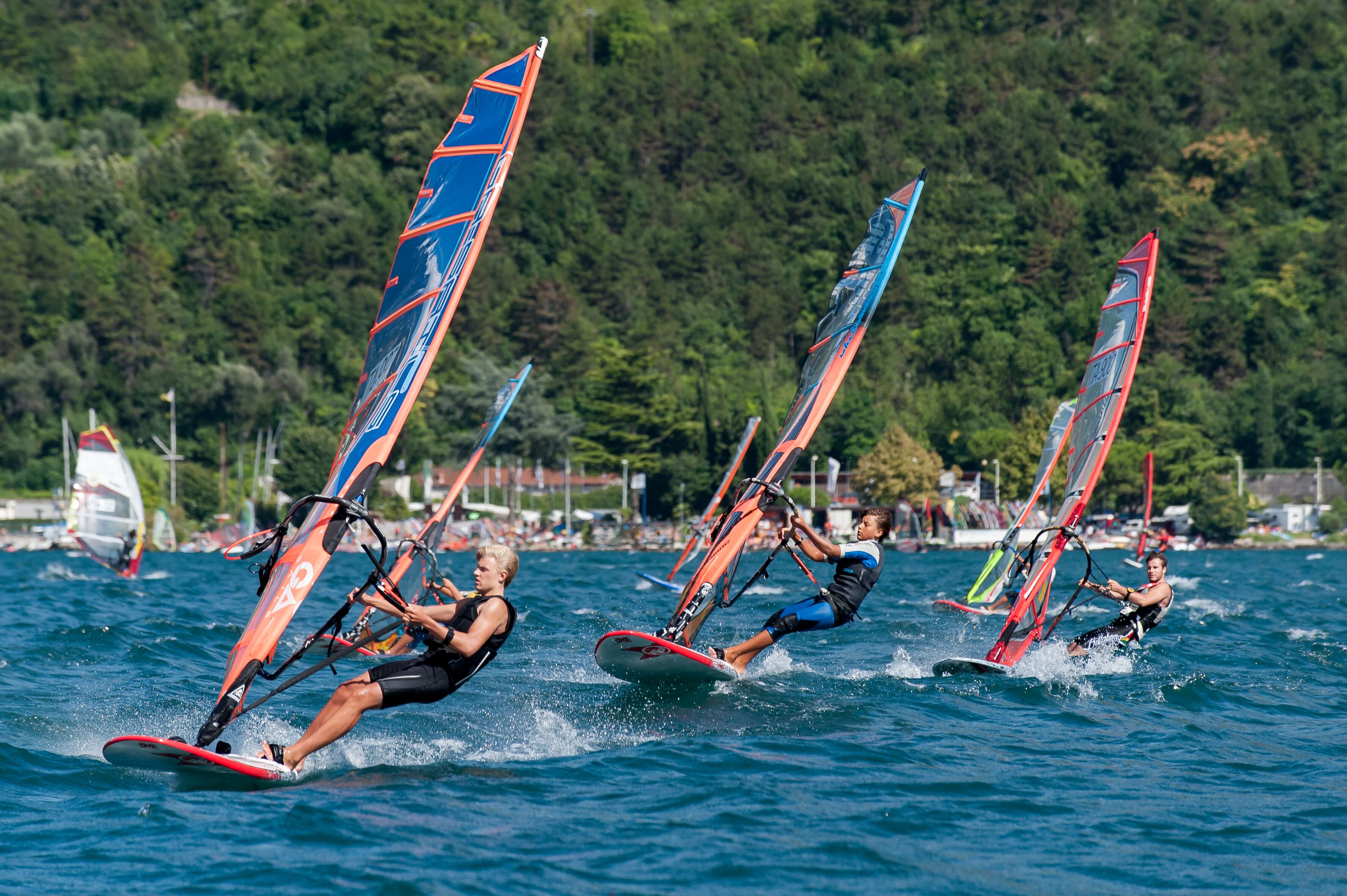 A group of windsurfing individuals on a body of water with a forested area in the background.