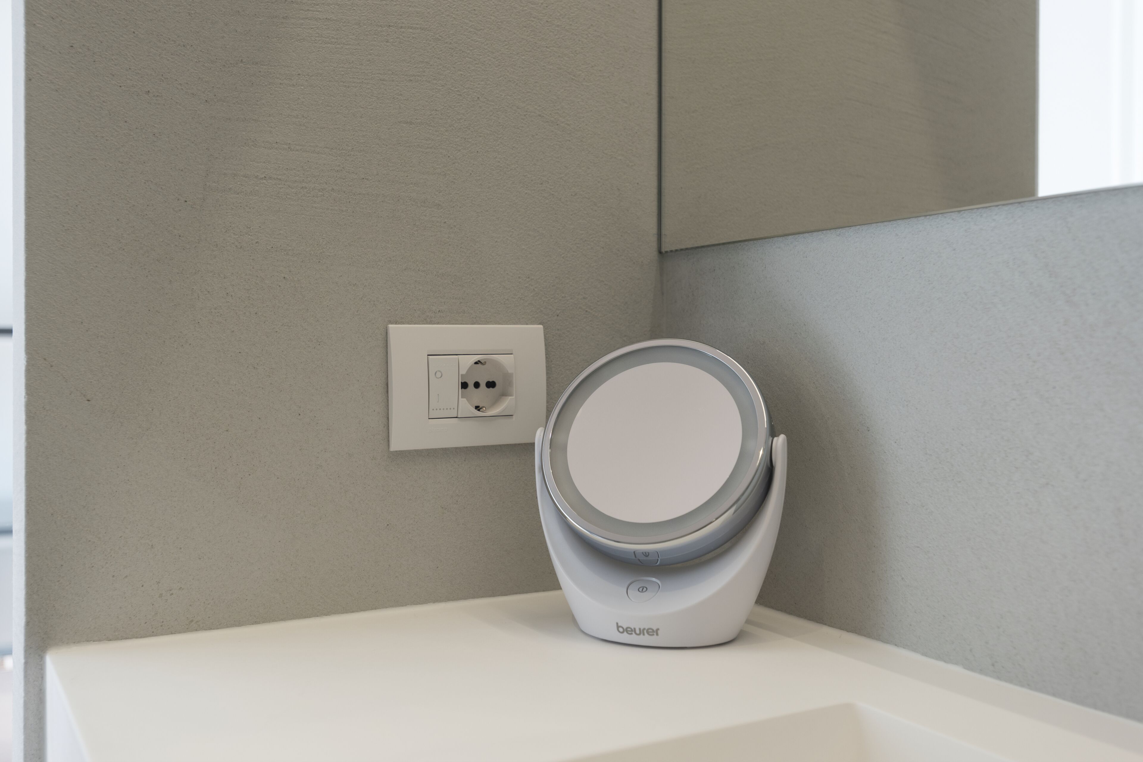 A modern bathroom with a white sink, a wall-mounted outlet, and a white, circular, contemporary mirror.