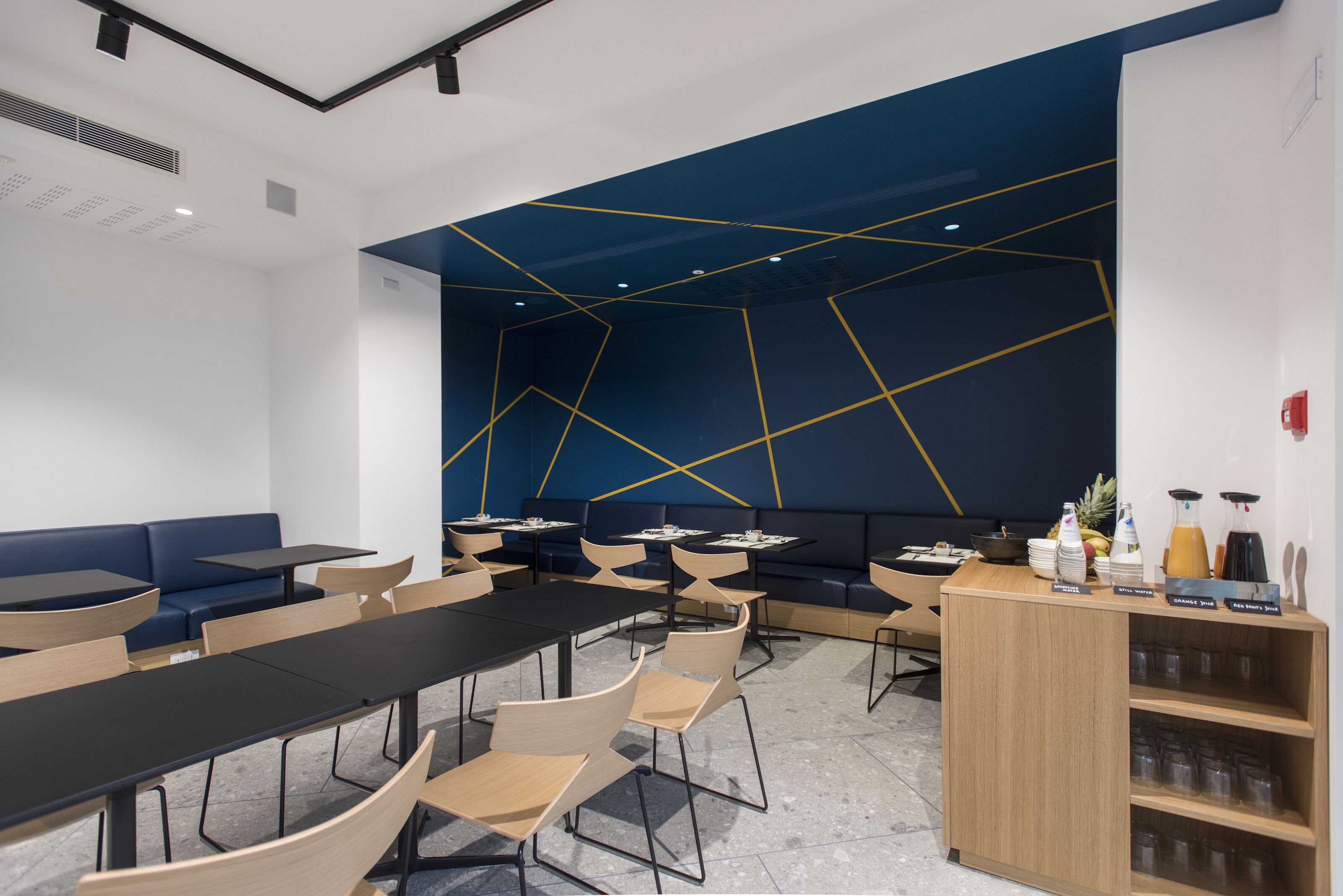 Une salle de conférence moderne dotée d’un mur au design géométrique, de tables noires et en bois, ainsi que d’un espace buffet proposant des boissons.