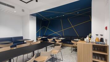 Une salle de conférence moderne dotée d’un mur au design géométrique, de tables noires et en bois, ainsi que d’un espace buffet proposant des boissons.