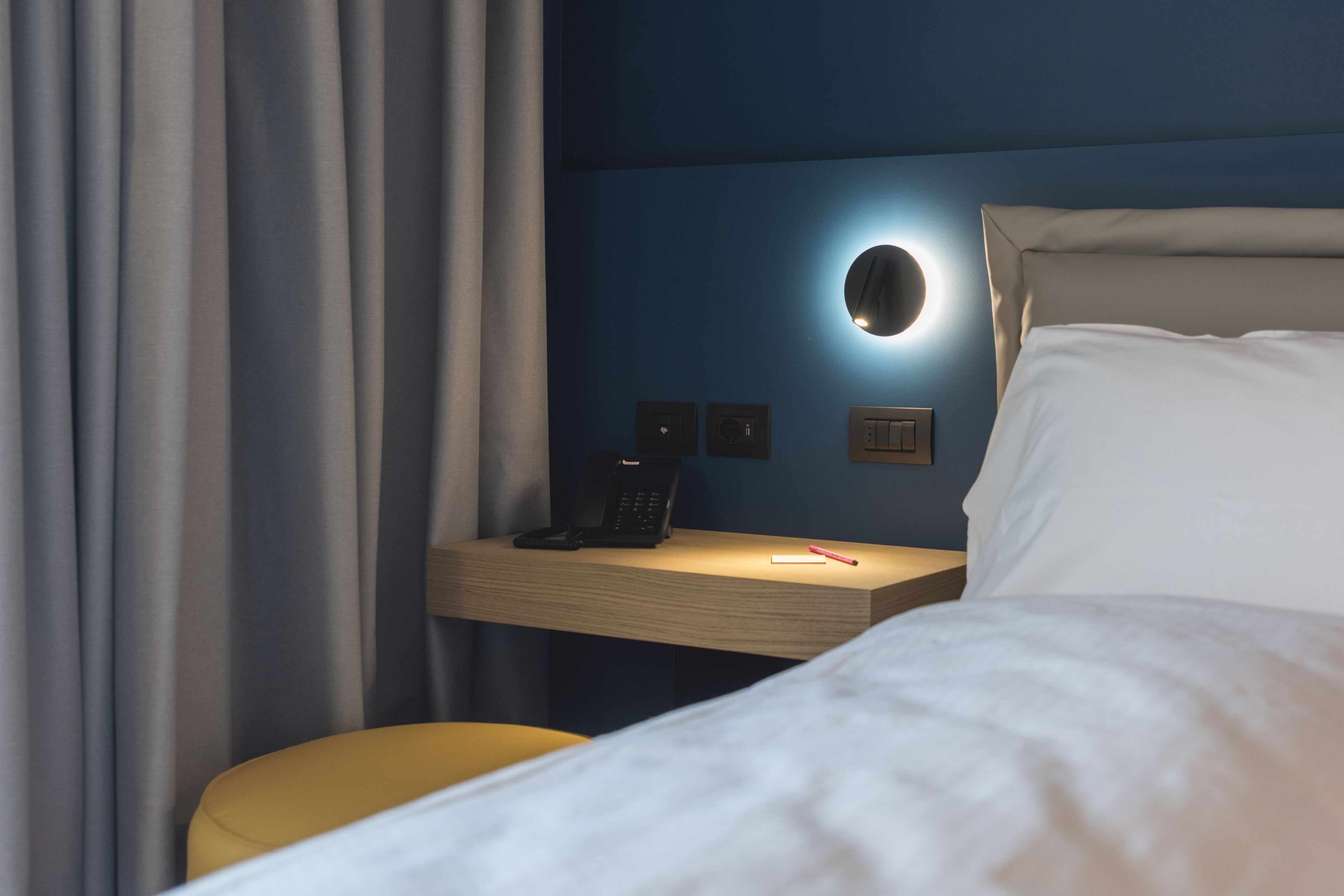 A hotel room with a bed, a bedside table with a phone, a lamp, and a chair.