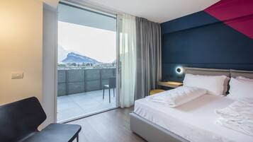 Habitación de hotel con cama, escritorio, silla, mesita de noche y vistas a la montaña desde un balcón.