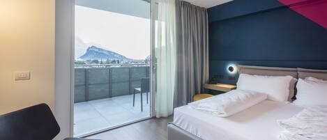 Habitación de hotel con cama, escritorio, silla, mesita de noche y vistas a la montaña desde un balcón.