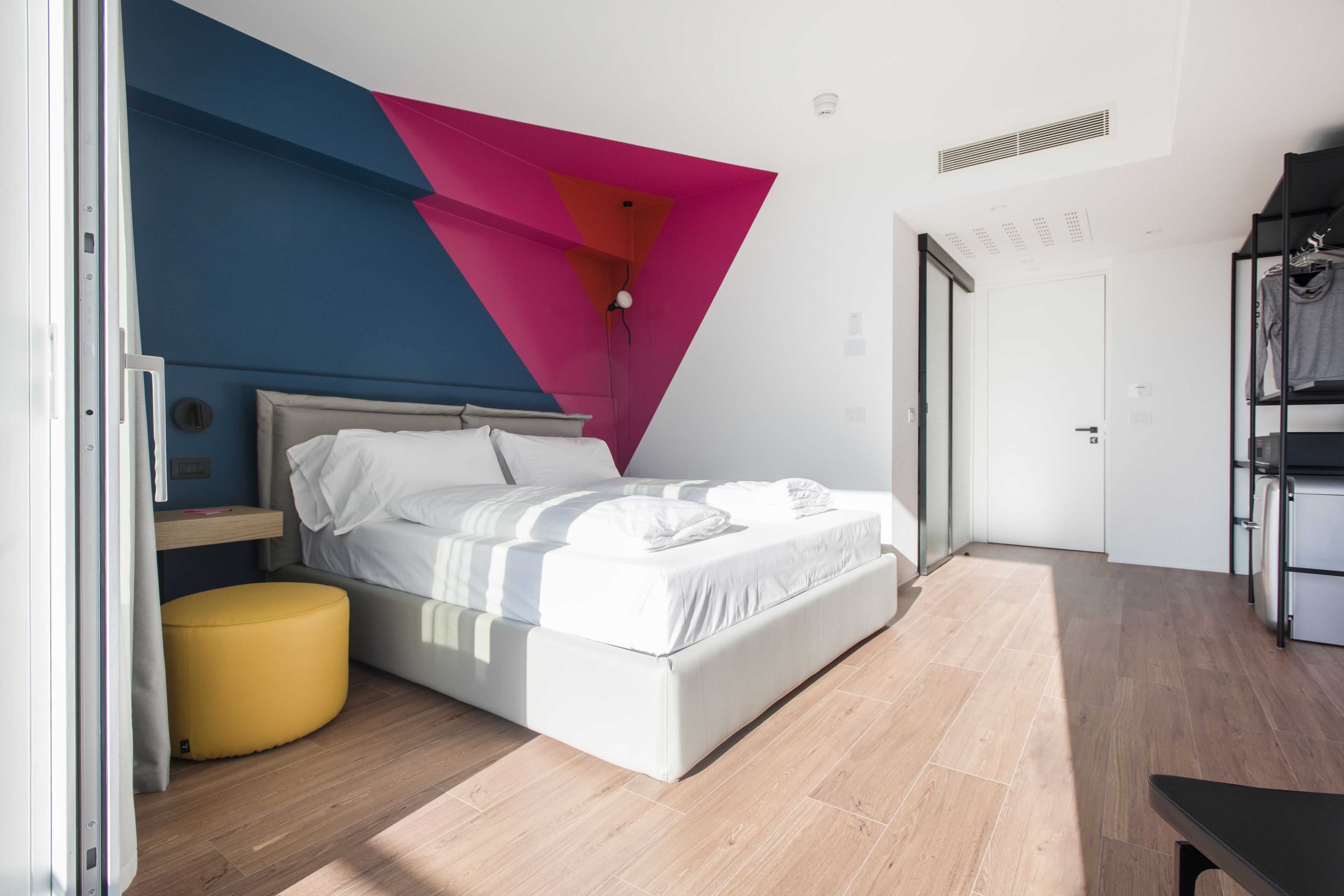 A modern bedroom with a bed, a yellow ottoman, and a unique geometric wall design.