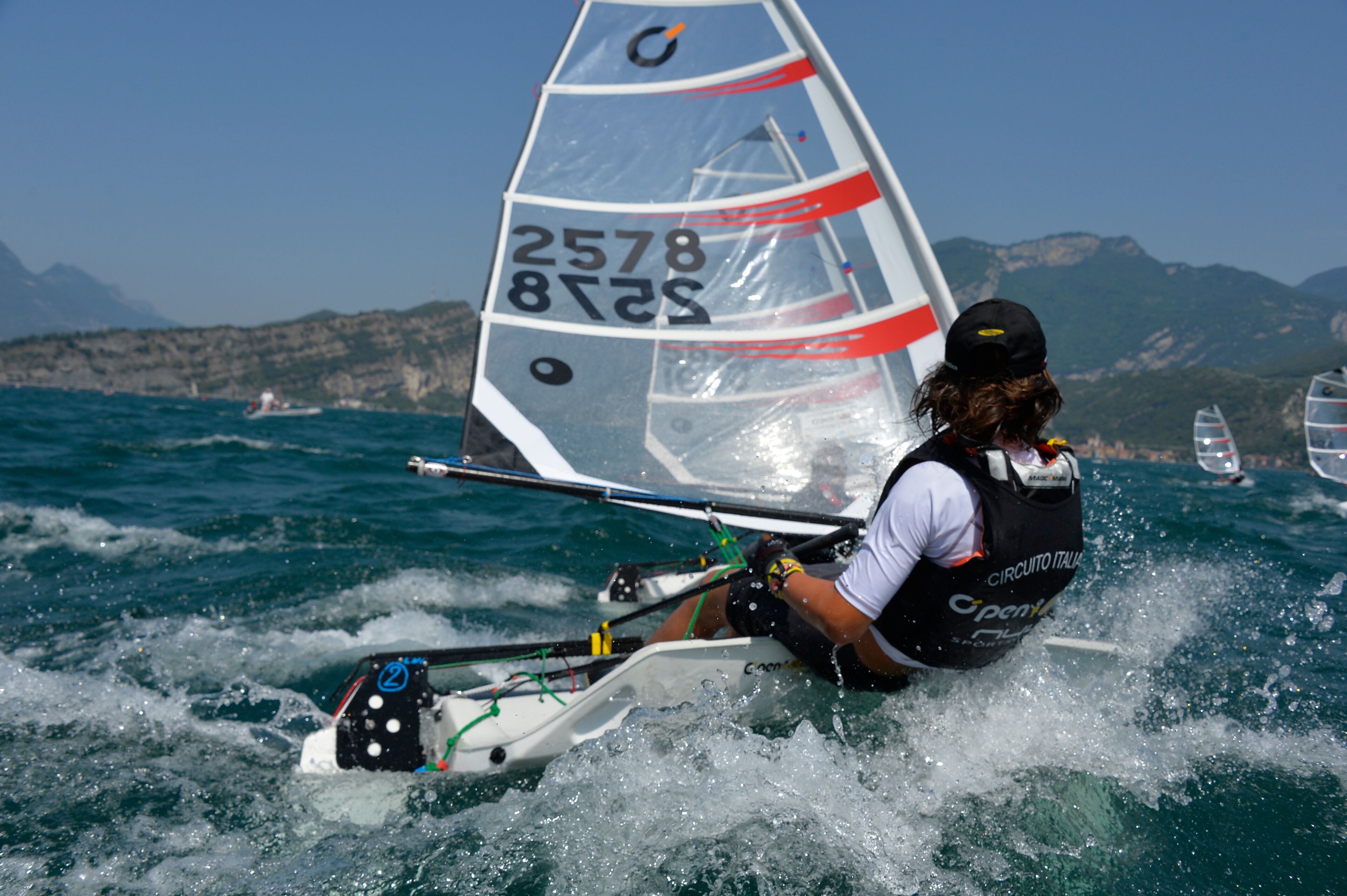 A person on a sailboat with a number 2578 sail, in a body of water with other boats and a mountainous coastline in the background.