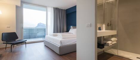 A modern hotel room with a bed, a chair, a bathroom with a glass shower, and a view of a mountain.