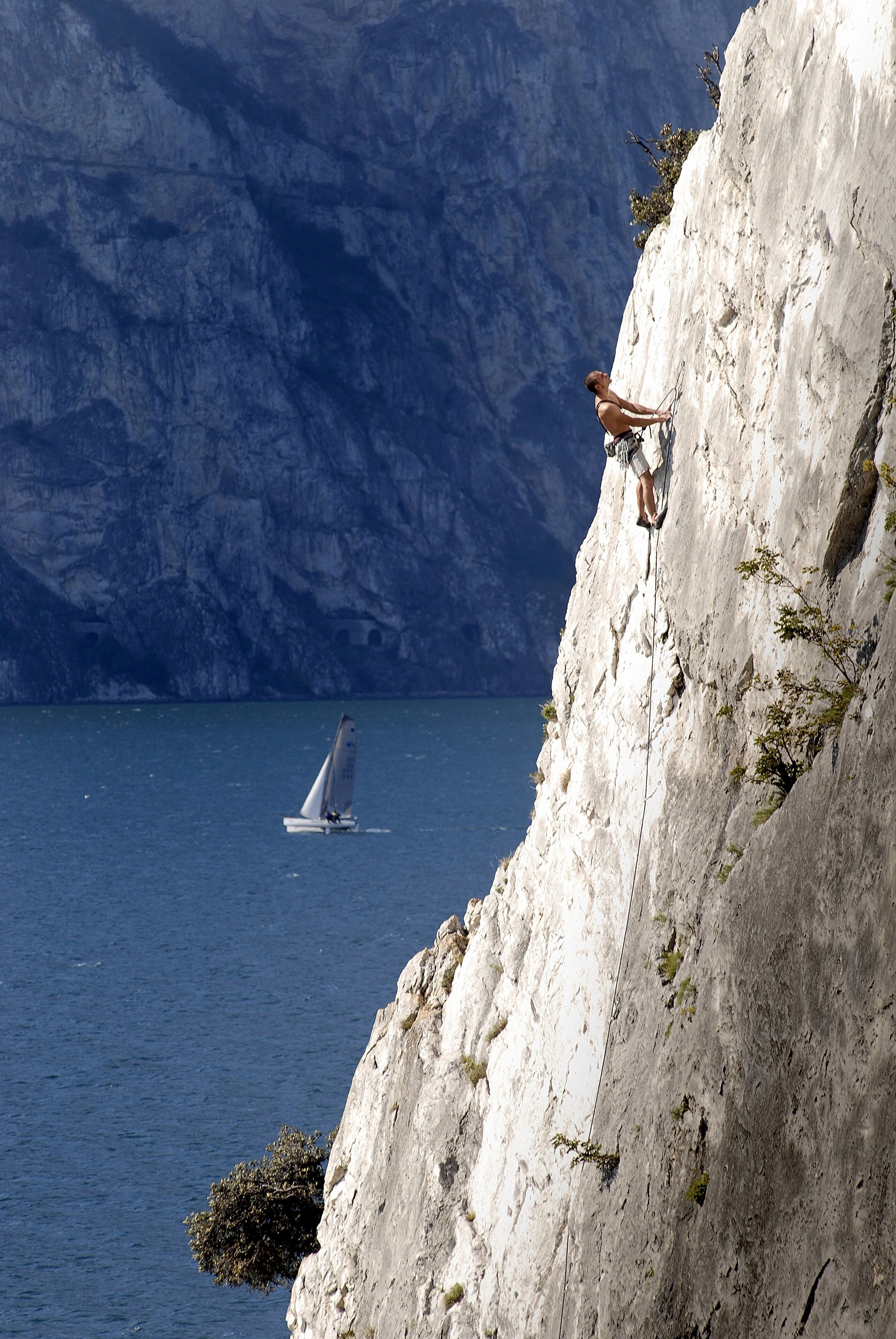 A person rappelling down a steep cliff with a sailboat in the background.