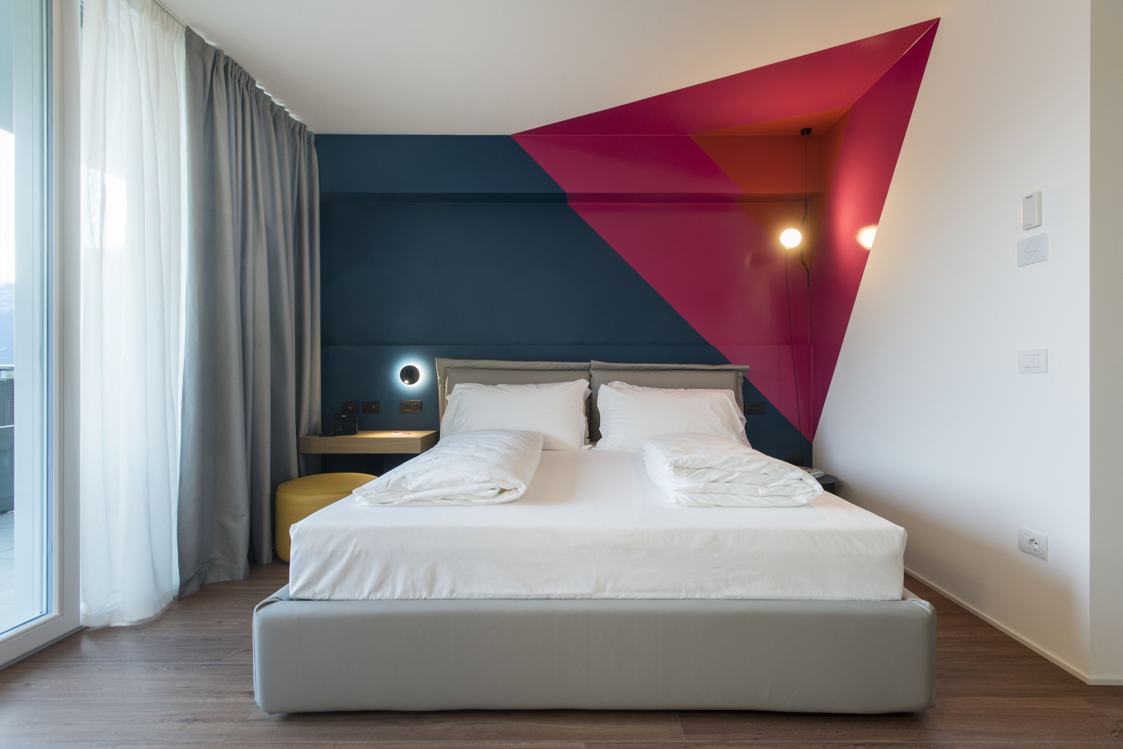Ein modernes Hotelzimmer mit einem großen Bett, einem Nachttisch mit Lampe und Blick ins Freie durch ein Fenster mit Vorhängen.