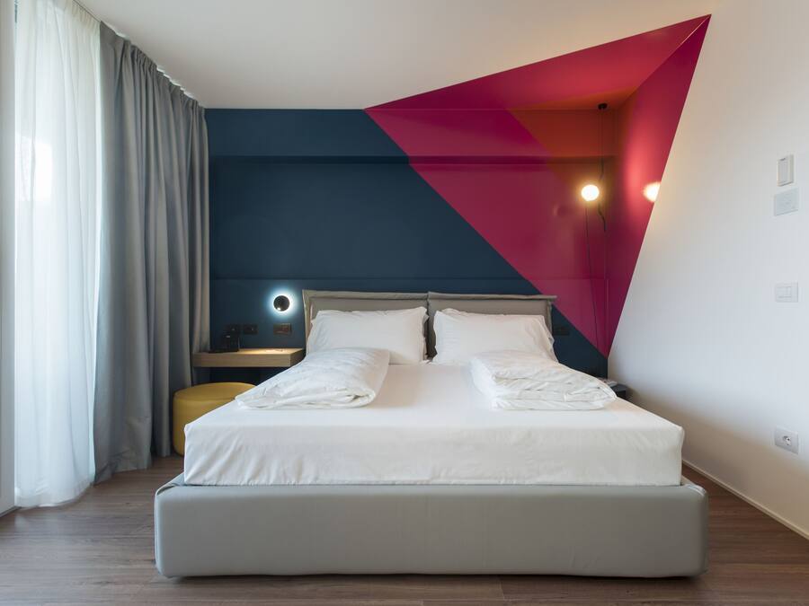 Habitación de hotel moderna con una cama grande, una mesita de noche con lámpara y vistas al exterior a través de una ventana con cortinas.