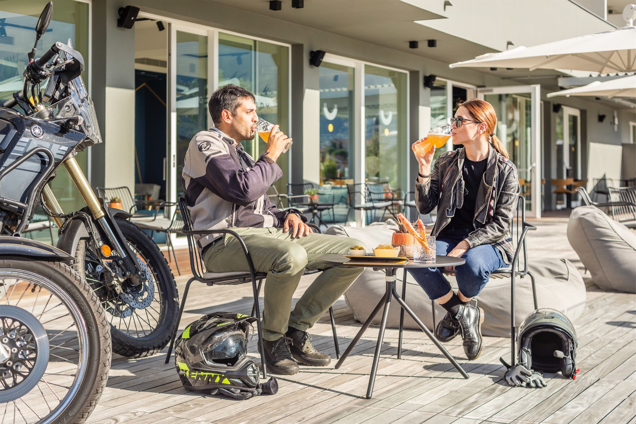 Two people sitting at an outdoor table with a motorcycle and a helmet nearby.