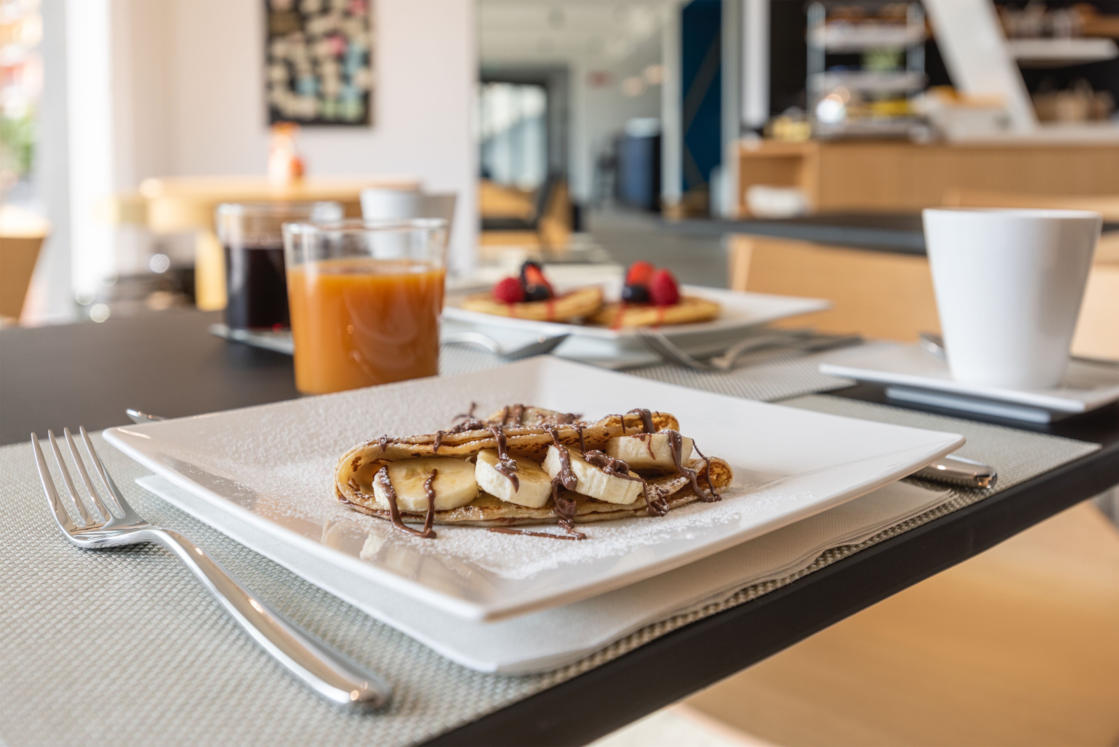 A breakfast setting with a plate of pancakes topped with bananas and chocolate, a cup of coffee, and a glass of orange juice on a table.