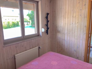 Een kamer met houten wanden, een roze beddeken, een radiator en een raam met uitzicht op groen en een gebouw.