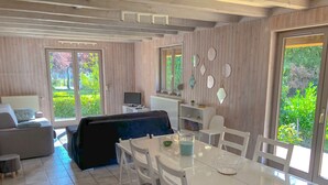 Un salon moderne comprenant un canapé, une table à manger avec des chaises, un téléviseur et une vue sur la verdure grâce à de grandes fenêtres.