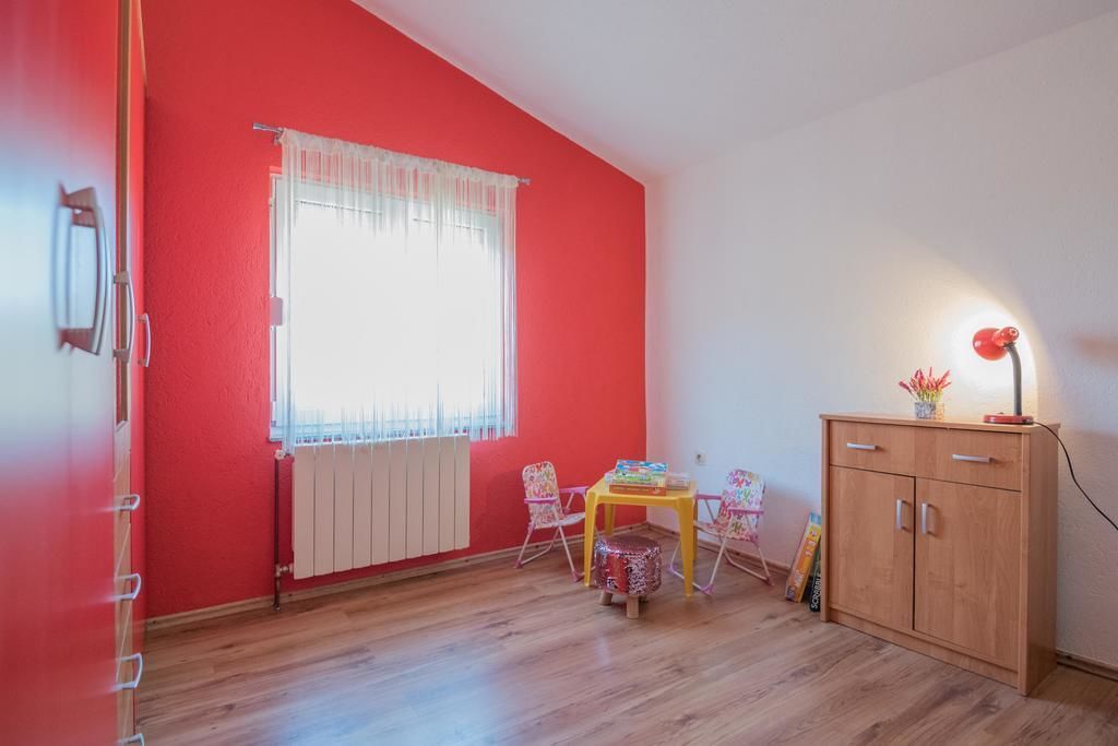 A room with a red wall, a window with white curtains, a wooden cabinet, a small round table, and two chairs.