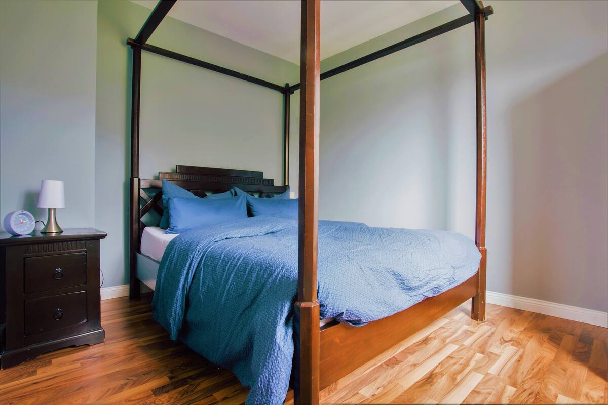 A four-poster bed with a blue bedspread, a nightstand with a lamp, and a wooden floor.