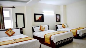 Habitación de hotel con tres camas, cada una con ropa blanca y almohadas decorativas anaranjadas. Hay cuadros enmarcados en las paredes, una ventana con cortinas y una puerta con espejo.