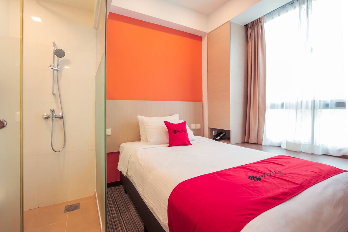 Habitación de hotel con ducha, cama con ropa de cama blanca y una manta roja, pared de acento naranja y ventana con cortinas.