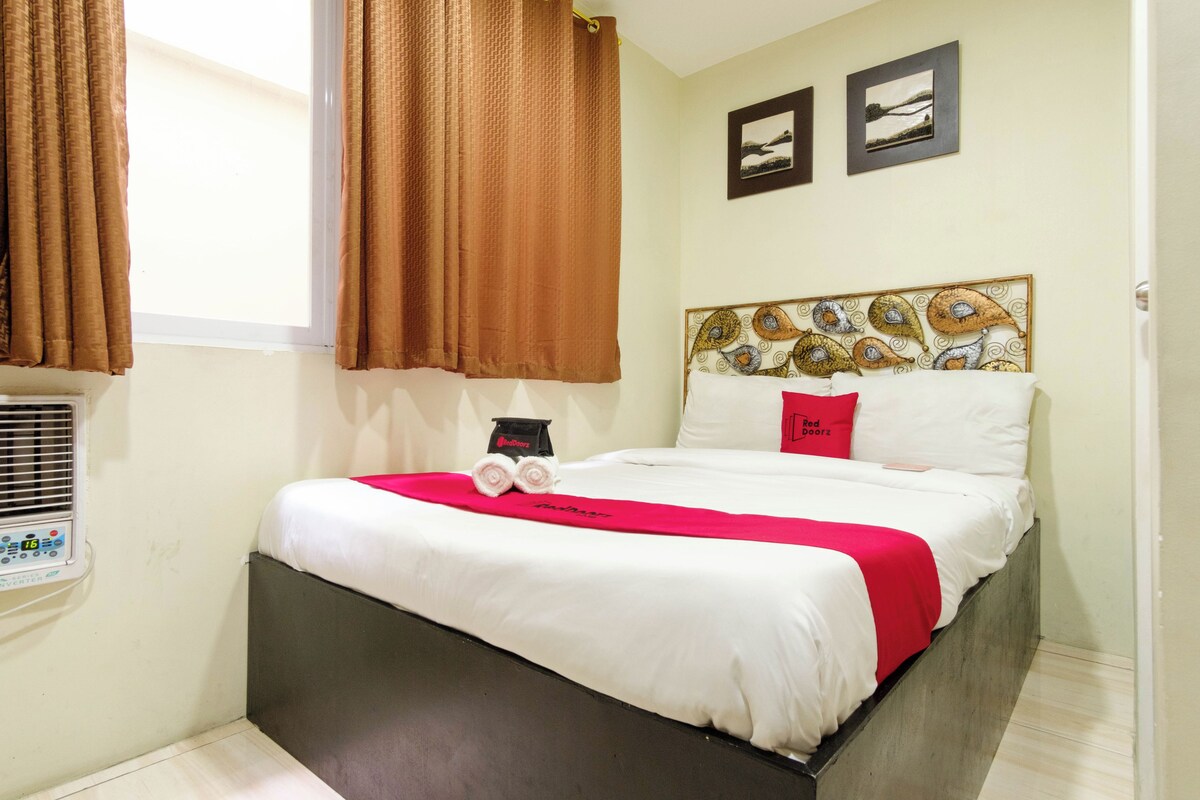 A neatly made bed with a red blanket and two pillows, a wall-mounted heater, and two framed pictures above the bed.
