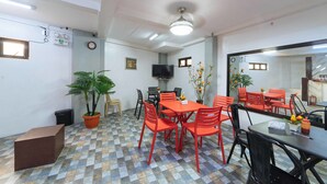 Un coin repas avec un sol à carreaux, une table centrale entourée de chaises, une plante en pot et un téléviseur fixé au mur.