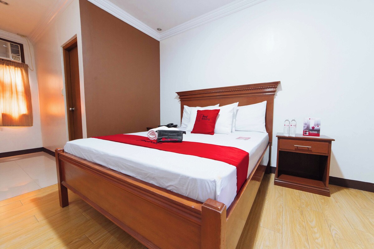 Un lit bien fait, avec un cadre en bois, une table de chevet dotée d’un tiroir et un coussin décoratif rouge.