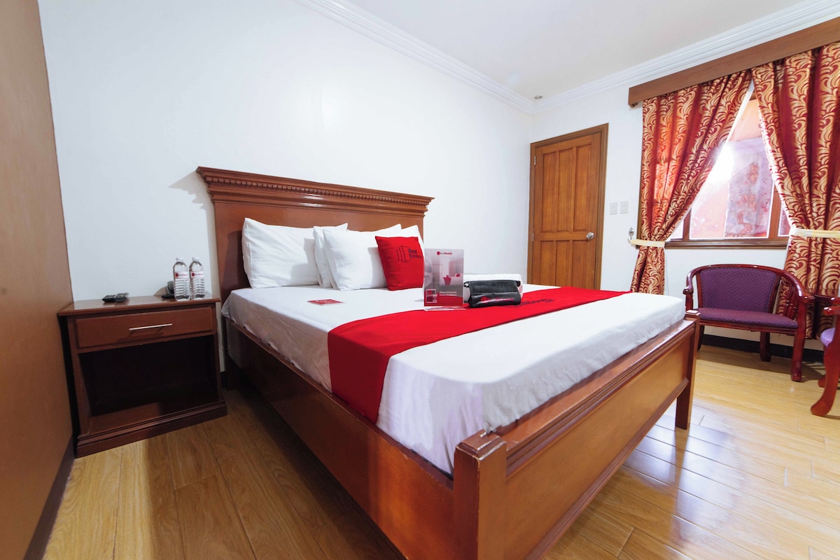 Une chambre d’hôtel avec un lit en bois, du linge de lit blanc, un plaid rouge, une table de chevet avec une lampe, une chaise et une fenêtre avec des rideaux.
