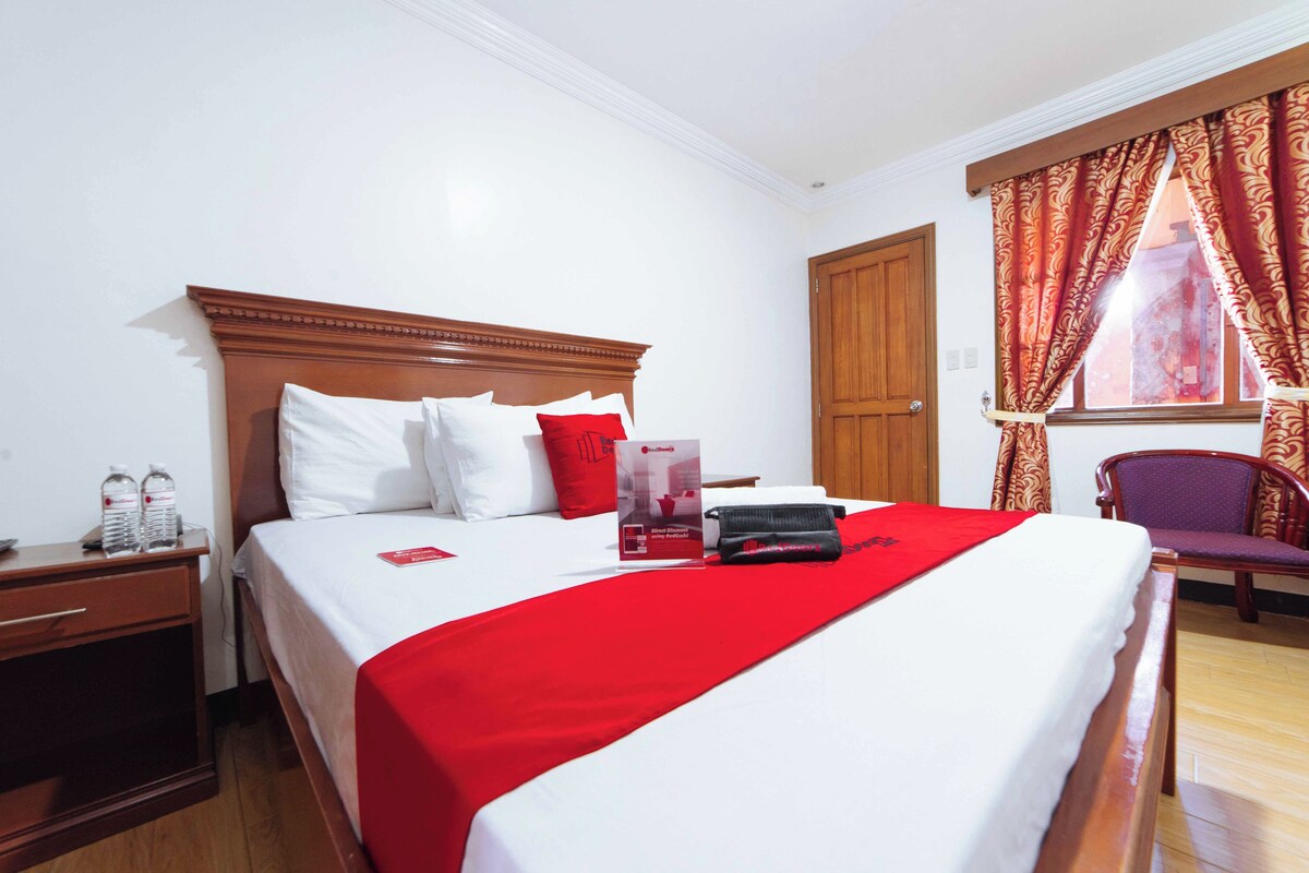 A hotel room with a wooden bed, white bedding, a red throw, a nightstand with bottles, and a chair by the window.