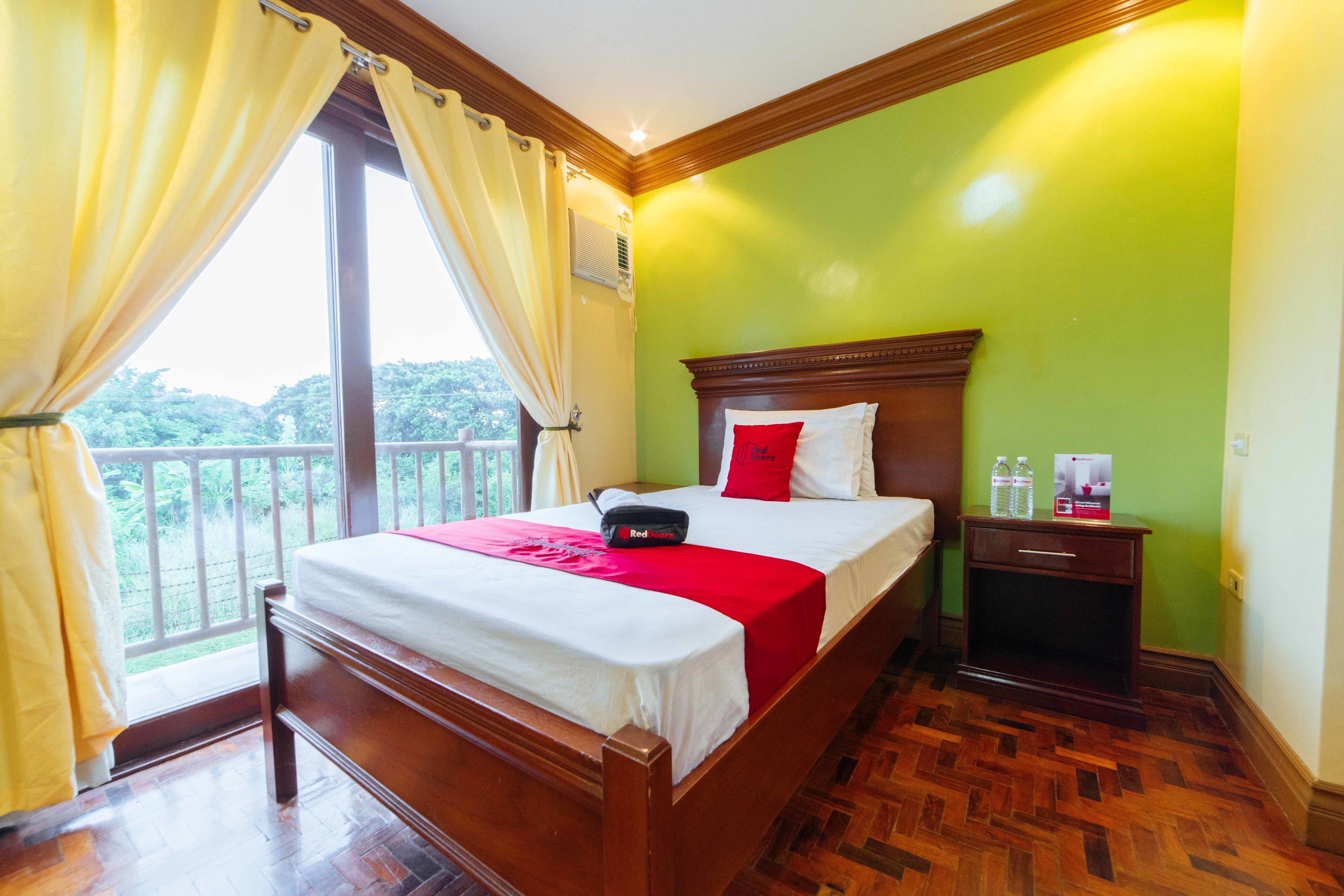 A hotel room with a wooden bed, a nightstand with a water bottle, and a view of greenery through the window.