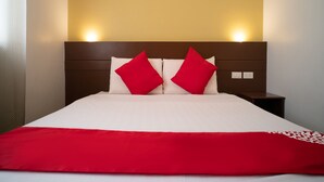 Una cama bien hecha con ropa blanca y dos almohadas rojas, cabecera de madera y dos lámparas fijadas a la pared.