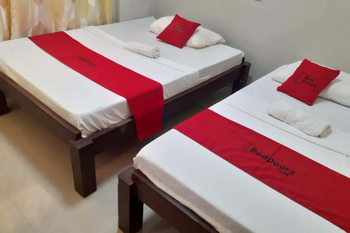 Two single beds with white linens and red accents, a wooden bench, and a wall-mounted light fixture.