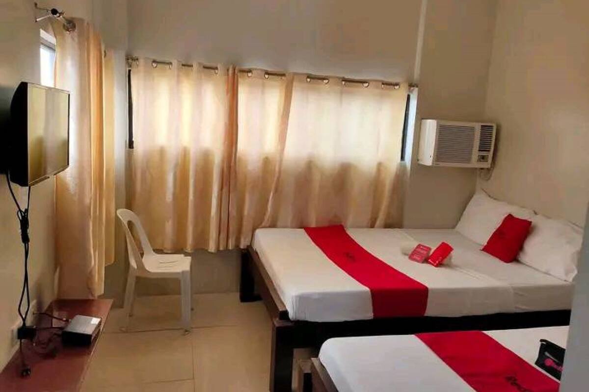 A hotel room with two beds, a desk, a chair, a TV, and an air conditioning unit.