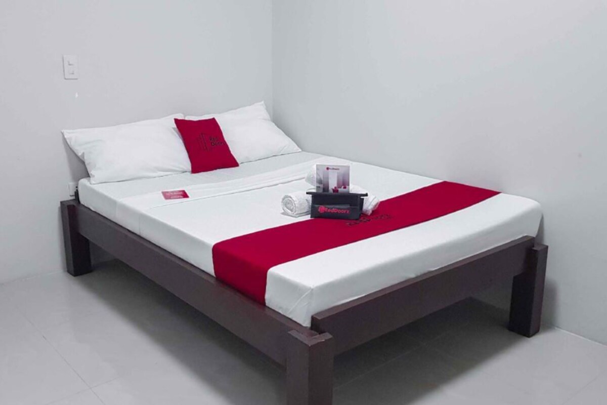 A single bed with a wooden frame, white bedding, a red accent pillow, and a bedside table with a tissue box and a small item.