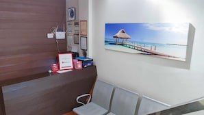 A reception area with a wooden desk, a computer, a clock, framed certificates, and a beach scene painting. - RedDoorz @ VP Inting Street (Tagbilaran)
