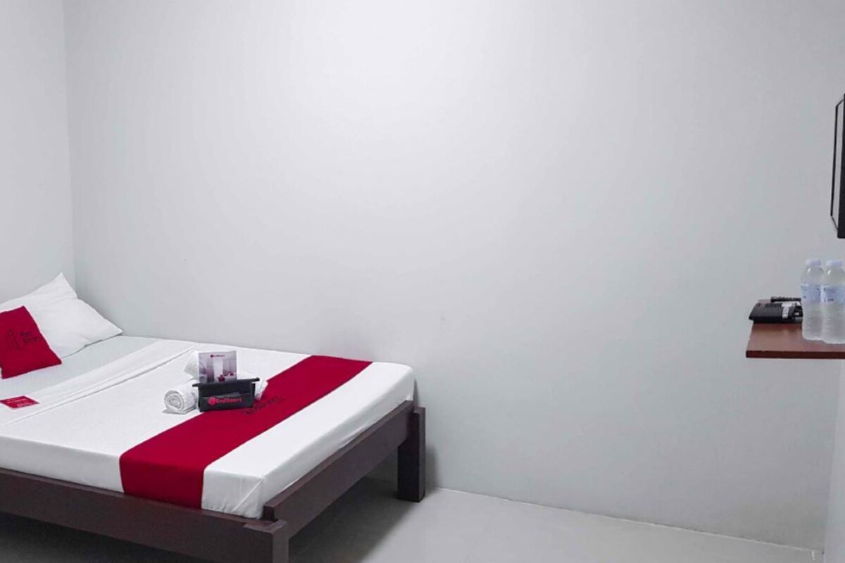 A single bed with a white and red bedspread, a red pillow, and a small bedside table with a water bottle and a remote control.