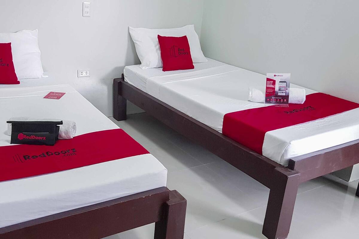 Two single beds with white bedding and red accents, a red blanket, a red pillow, a black object, and a RedDoorz branded box.