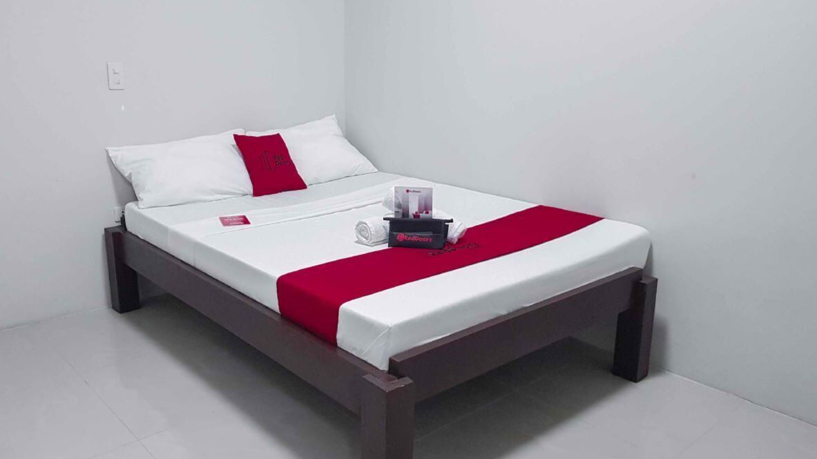 Uma cama de solteiro com estrutura de madeira, roupa de cama branca, um travesseiro decorativo vermelho e uma mesa de cabeceira com um estojo de lenços e um pequeno objeto.