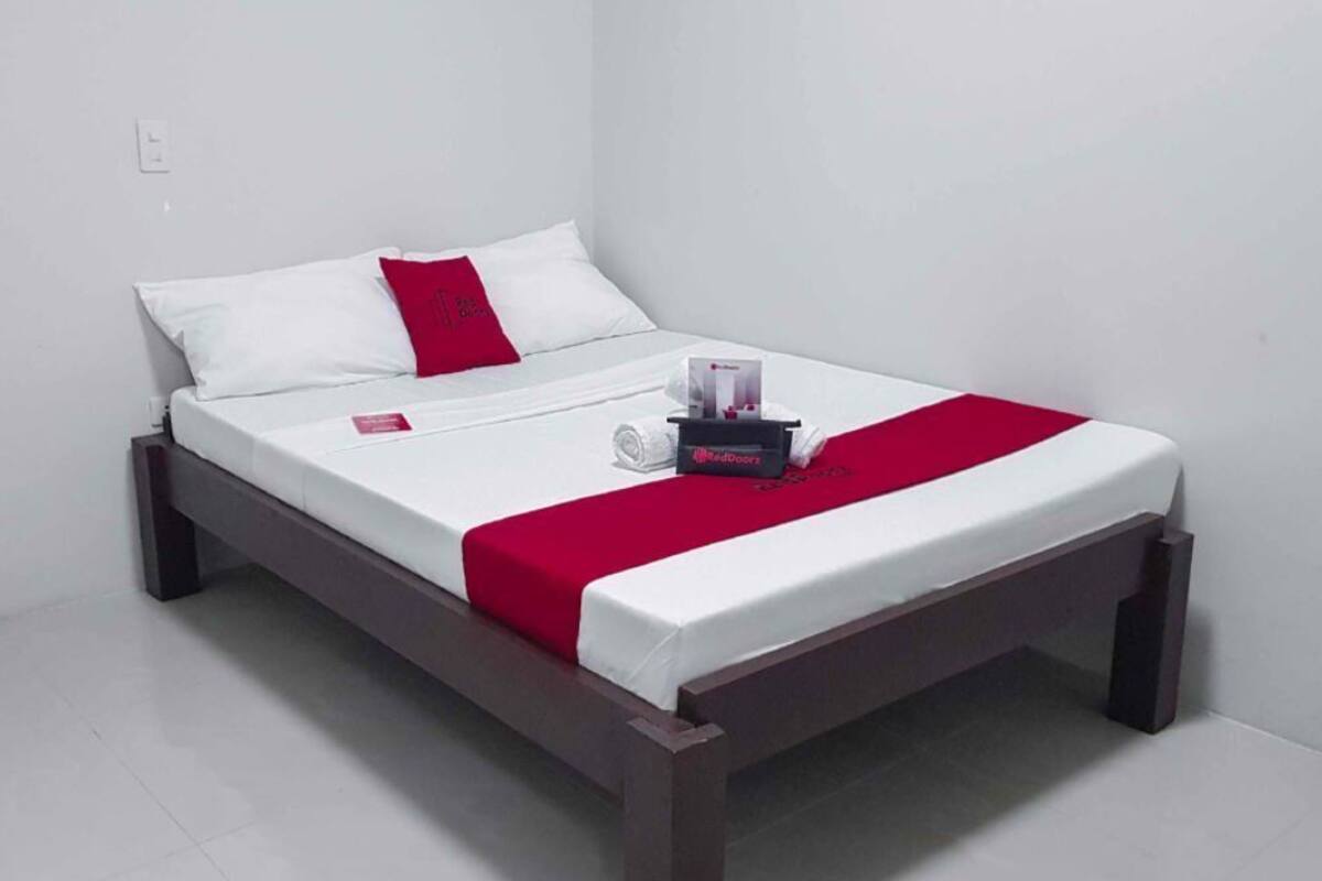 A single bed with a wooden frame, white bedding, a red accent pillow, and a bedside table with a tissue box and a small item.