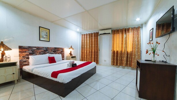 A hotel room with a large bed, a wooden headboard, a bedside table with a lamp, a dresser, a TV mounted on the wall, and a window with curtains. - RedDoorz Premium @ Clarkview Angeles City (Angeles City)