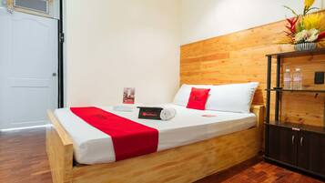 A neatly made bed with a red and white bedspread, a RedDoorz towel, and a small bedside table with a bottle and a tissue box.