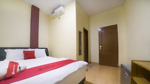 Une chambre d’hôtel avec un lit, une couvre-lit rouge, un climatiseur fixé au mur, une porte en bois et une petite table en bois.