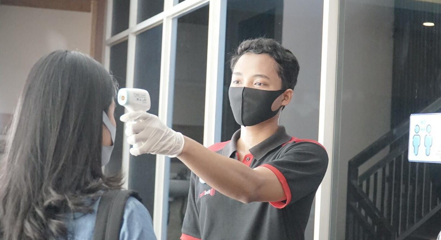 Une personne portant un t-shirt noir et rouge et un masque facial, tenant un appareil près du visage d’une autre personne.