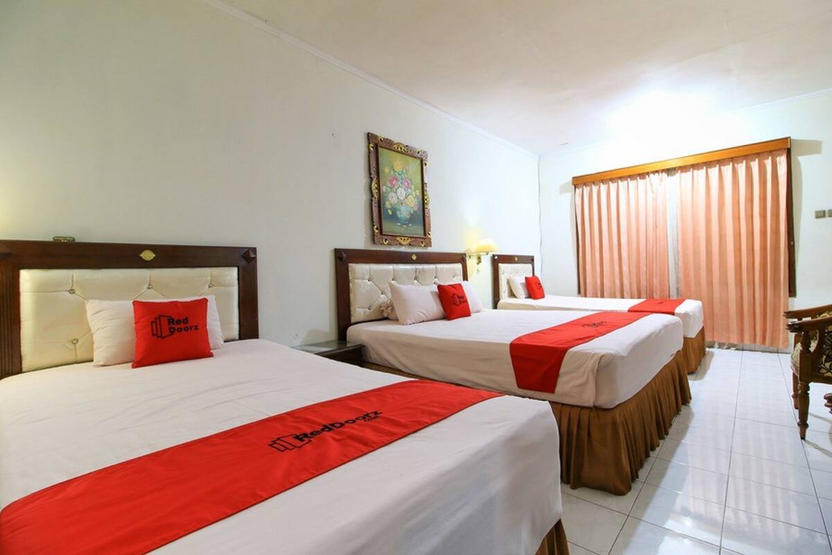 Een hotelkamer met drie bedden, elk voorzien van een rode sprei en kussens, een houten hoofdbord, en een ingelijst schilderij aan de muur.